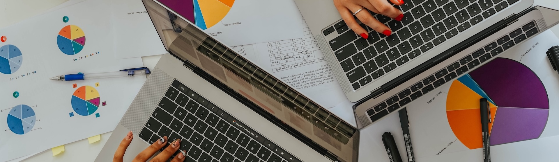 Two members of a marketing agency type on the keyboards of their laptops on a desk filled with pie charts outlining campaign growth and performance